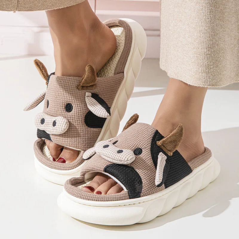 Tan knit cow slippers with embroidered cow face, brown ears, and plush strap detail.